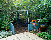 Kinderschaukel mit Rindenspänen, blauem Spalier, Feige (Ficus Brown Turkey), Fatsia Japonica und Terrakotta-Wasserspiel