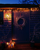 THE Blue SUMMERHOUSE at NIGHT with Silver WREATH, TWISTED WILLOW TREES IN Purple PLASTIC Container, CANDLE AND FAIRY LIGHTS