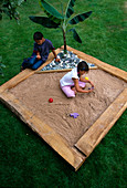 Designer: Clare MATTHEWS - Tropical Island SANDPIT - WOODEN FRAMEWORK with CROSS FRAME AND PLASTIC SHEETING, Banana, PEBBLES, Sand, Girl AND Boy