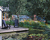 VIEW ACROSS LILY Pool TO DECK with Adirondack CHAIRS. IN THE BACKGROUND IS THE Main GREENHOUSE. GREYSTONE Cottage, OXFORDSHIRE. Designer: DUNCAN HEATHER