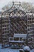 Metal arbour with snowy bench in front of a hedge of Carpinus (hornbeam)