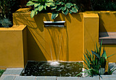 Wasserspiel: 'Briefkasten'-Wasserfall mit gelbgetrichenen Wänden und Hosta im Hintergrund
