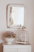 Vintage birdcage an urn of flowers on white cabinet below mirror on wall