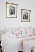 Sofa mit weisser Husse und nostalgischen Kissen, darüber gerahmte schwarz-weiße Fotos