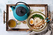 Bowl of asian style soup with noodles, egg, spring onion and tofu cheese, teapot and cup of green tea