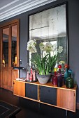 Colourful glass bottles and bowl of white-flowering plants on retro sideboard below mirror on wall