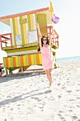 A brunette woman wearing a pink dress, statement necklace and sneakers in front of a lifeguard tower on the beach