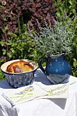 Hand-made pot holders, cake in ceramic pot and lavender in ceramic jug on white tablecloth on small garden table