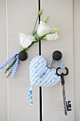 Pale blue, ornate, heart-shaped pendant and key next to white artificial flowers on cabinet door knobs