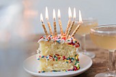 Geburtstagskuchen mit brennenden Kerzen und bunten Zuckerstreuseln neben Champagnerglas