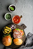 Fresh homemade burgers on dark serving board with spicy tomato sauce, green salt, pepper, pickles and onion