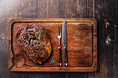 Grilled Black Angus Steak Ribeye and fork and knife on meat cutting board on dark wooden background