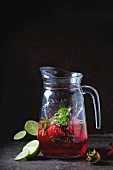 Glass jug of strawberry lemonade, served with fresh strawberries, mint and lime over dark background