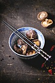 Porcelain bowl of dry shiitake mushrooms and black chopsticks over dark table