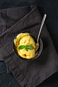 Homemade mango ice cream with fresh mint in vintage iron bowl on dark textile napkin over black background