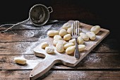 Uncooked homemade potato gnocchi with fork and strainer on vintage cutting board over wooden table with flour