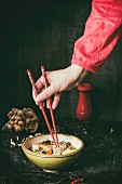 Female hand in red shirt take by chopsticks noodles from asian ramen soup with shrimp, onion, sliced egg and mushrooms