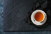 White vintage cup of hot tea on saucer standing on black textile napkin over black slate texture background