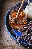 Open glass jar of liquid honey with honeycomb and honey dipper inside, milk and dry lavender