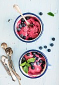 Homemade blueberry ice cream scoops with fresh berries and mint leaves in enamel mugs over light blue background