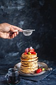Dusting Icing Sugar on Stack of Gluten free Oat Flour Pancakes served with chopped Strawberries and Maple Syrup