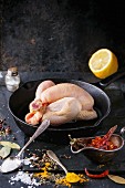 Raw mini Chicken in Cast-iron pan with sea salt and different spices over black slate board