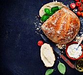 Selbstgebackenes Brot umgeben von Sandwich-Zutaten wie Tomaten, Basilikum, Öl und Frischkäse