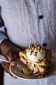 A man is holding a slice of a caramel pie topped with vanilla ice cream and chopped walnuts
