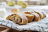 Sliced fresh baked homemade apple strudel over towel on kitchen table with jug of milk and apples