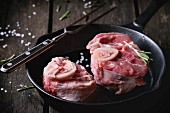 Raw osso buco meat in cast-iron pan with vintage meat fork, salt, pepper and rosemary over old wooden table