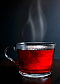 Hot red berry fruit tea in a glass mug with steam standing on a dark wood with dark grey background