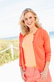 A blonde woman on the beach wearing a T-shirt, cardigan and trousers in different orange tones