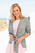 A blonde woman wearing a pink dress and grey cardigan on the beach