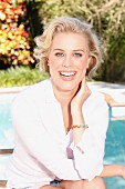 A blonde woman wearing a white blouse and shorts by a pool