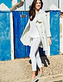 A brunette woman wearing a white trench coat, a lace top, jeans with fringing on the seam and perforated slip-on shoes
