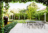 Terrace area with table and chairs made of metal, surrounded by green plants and lawn