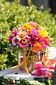 Farbenfroher Blumenstrauß mit Zinnias und Tagetes in Glasvase auf Vintage Gartenstuhl