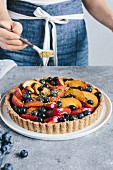A woman is sprinkling a peach and blueberry tart with pistachios