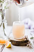 Pink Grapefruit Cocktail Pouring into Glass, with Flowers on White Wood