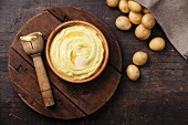 Wooden bowl with mashed potatoes and raw potatoes on dark background