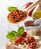 Tomato pesto and basil leaves on a wooden spoon above a wooden board with basil and dried tomatoes