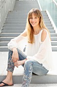 A blonde woman wearing a white blouse, jeans with embroidered flowers and flip flops, sitting on a flight of stairs