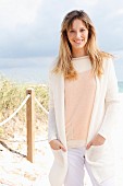 A blonde woman wearing a cream jumper, a white cardigan and white trousers on the beach