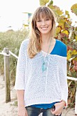 Blonde Frau in blauem T-Shirt, weißem Strickpullover und Jeans am Strand