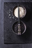 Schwarzer und weisser Reis in Schälchen mit Stäbchen auf schwarzem Untergrund