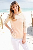 A blonde woman wearing a cream jumper and white trousers on the beach