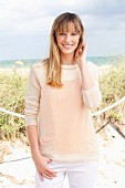 A blonde woman wearing a cream jumper and white trousers on the beach