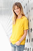 A blonde woman wearing a yellow T-shirt and jeans in front of a wall with holes