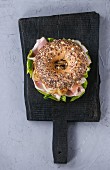 Whole Grain bagel with fried onion, green salad and prosciutto ham on black wood choping board