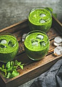 Fresh green smoothie in glasses with ice cubes, mint and lime in wooden tray over grey concrete background